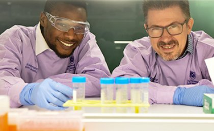 two men in purple lab coats and wearing glasses look at a rack of blue-lidded sample pots